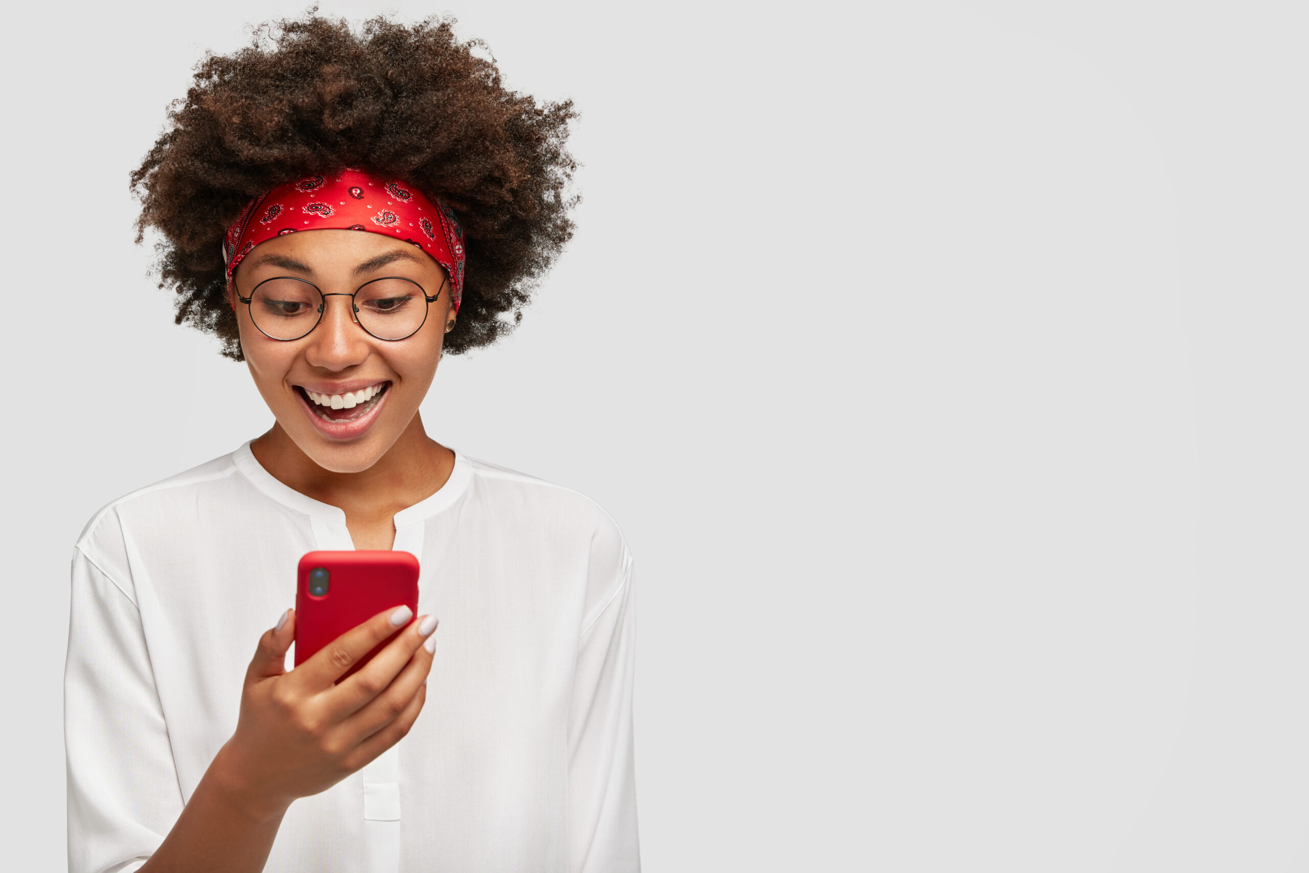 Horizontal shot of pleasant looking black adult, excited with video chat, carries up to date cell phone, has broad smile, wears fashionable red headband, models in studio against blank copy space